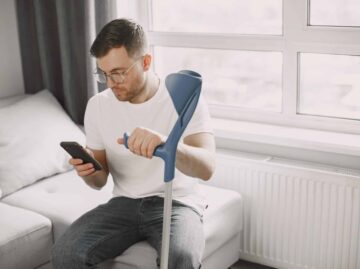 Young man with walking stick talk by phone indoors Startseite young man with broken leg home using smartphone scaled scaled Versicherungspapa